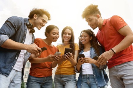 Foto de Grupo de jóvenes multiétnicos con teléfonos inteligentes en las manos de pie al aire libre, estudiantes felices mirando a la pantalla del teléfono móvil con interés, Comprobación de la nueva aplicación educativa o sitio web, Primer plano Shot - Imagen libre de derechos