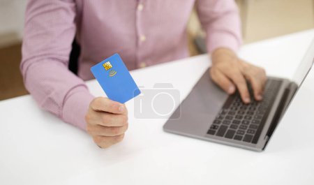 Photo for Caucasian mature guy at table with laptop uses credit card in office interior. Saving money recommendation, finance, businessman and business, manager work, online shopping and sale - Royalty Free Image