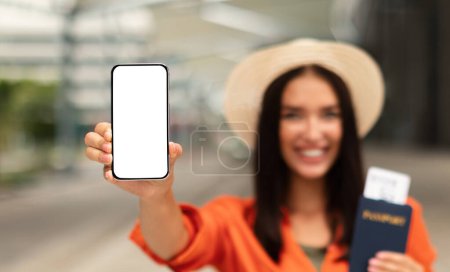 Foto de Viaje inteligente con tecnología móvil. Happy Lady Mostrando la pantalla del teléfono en blanco sosteniendo la tarjeta de embarque en el pasaporte, promoviendo la aplicación de viajes para reservar boletos en línea al aire libre. Burla, enfoque selectivo - Imagen libre de derechos