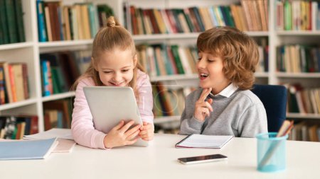 Foto de Dos niños de la escuela primaria sentados juntos en el escritorio en el interior del aula o de la biblioteca, usando una tableta digital, jugando juegos o viendo videos educativos durante el descanso - Imagen libre de derechos