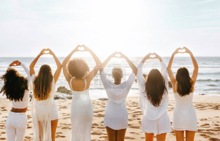 Foto de Vista posterior de las mujeres jóvenes haciendo símbolo del hogar con las manos, de pie en la línea de la orilla del océano al atardecer, espacio libre. Diversas damas celebrando despedida de soltera en la costa - Imagen libre de derechos