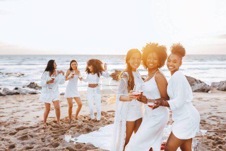 Téléchargez les photos : Dames multiraciales excitées posant avec des verres à vin tout en célébrant la fête des poules sur la plage au bord de l'océan, en s'amusant à la côte et en profitant de leurs vacances - en image libre de droit