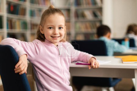 Photo for Portrait of pretty smart female elementary school pupil, sitting at desk in classroom, turning back and smiling at camera during lesson in school, free space - Royalty Free Image