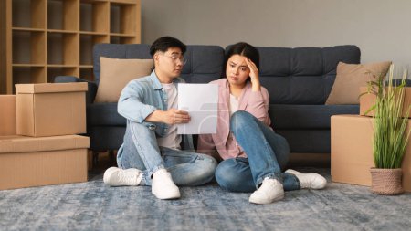 Photo for Home Loan Problem. Unhappy Asian Family Couple Having Issue With Mortgage Debt, Holding Papers Sitting Among Carton Moving Boxes Indoor. Apartment Rental Documentation Problems. Panorama - Royalty Free Image