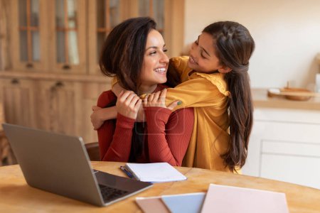 Foto de Happy Arab Little Daughter Abrazando a mamá mientras trabaja en línea en el ordenador portátil, sentado en el escritorio en casa. Madre y niña acurrucándose, posando cerca de la computadora. Tecnología de Internet y vida familiar - Imagen libre de derechos