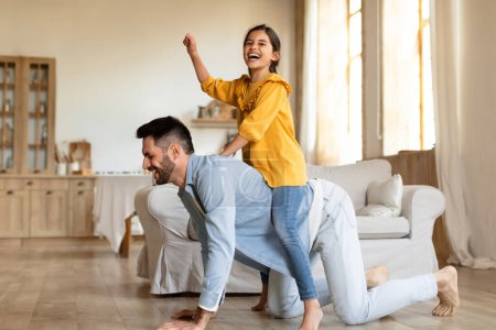 Photo for Family fun and games. Happy father playing with his little daughter, piggybacking girl, giving ride on his back, joyful kid playing cowgirl on horse at modern living room at home interior - Royalty Free Image