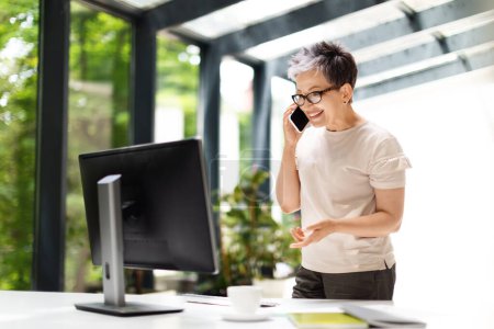 Photo for Positive successful attractive mature woman wearing casual outfit and eyeglasses CEO manager working at green office. Lady entrepreneur have phone conversation, looking at computer pc screen - Royalty Free Image