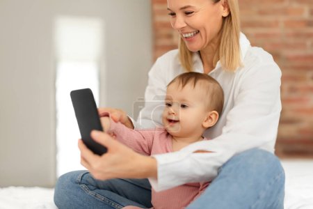 Photo for Happy european mother and infant baby girl using mobile phone playing educational games online or watching cartoons, sitting on bed. Baby and gadget use concept - Royalty Free Image