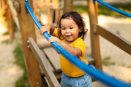 Foto de Adorable niña asiática jugando sola en el patio de recreo afuera, parada en el puente oscilante al aire libre, divirtiéndose en el parque de atracciones en el día de verano. Concepto de juego y ocio infantil - Imagen libre de derechos