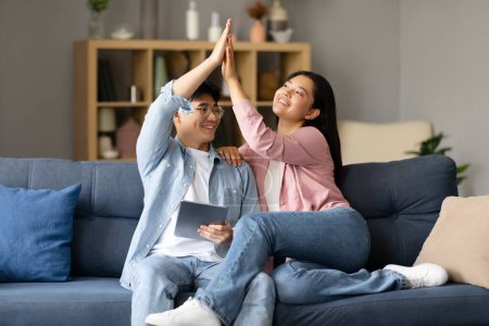 Téléchargez les photos : Couple japonais joyeux utilisant la tablette numérique et donnant haute cinq assis à la maison. Jeune copine et petit ami se détendre avec ordinateur célébrer le succès en ligne. Gadgets et mode de vie moderne - en image libre de droit