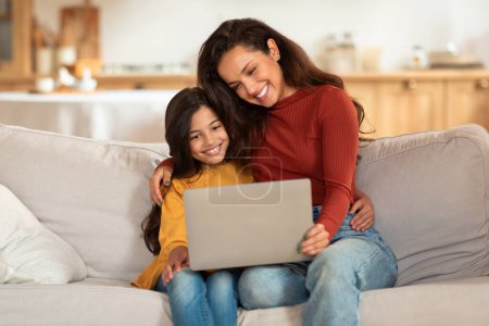 Foto de Feliz joven árabe madre e hija usando ordenador portátil, Websurf y viendo dibujos animados en línea juntos sentados en el sofá en casa. Mamá y niño chica navegar por Internet a través de la computadora - Imagen libre de derechos
