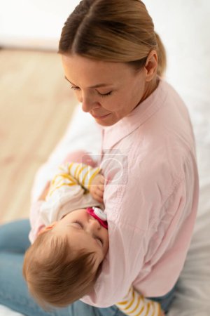 Photo for Loving mother holding her cute sleeping infant daughter on hands, comforting, lulling kid and looking at her, sitting on bed in bedroom at home, above view, vertical shot - Royalty Free Image