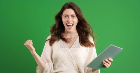 Photo for Glad excited european millennial woman using tablet, doing success gesture, celebrating victory, isolated on green studio background. Good news, ad and offer, huge sale, win emotions - Royalty Free Image