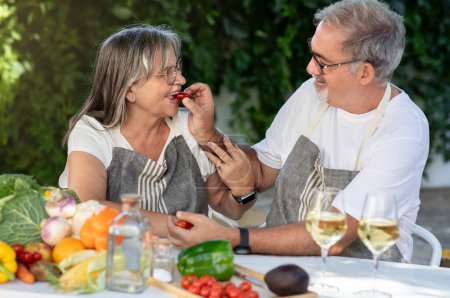 Téléchargez les photos : Joyeux vieux mari européen dans tablier nourrir femme avec des tomates à table avec des légumes biologiques, de plein air. Repas sains à la maison, cuisine ensemble, déjeuner romantique, dîner dans le jardin - en image libre de droit