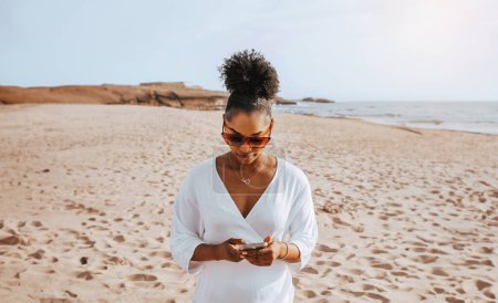 Foto de Mujer negra viajera vestida de blanco usando celular en la playa, parada en la costa de la ciudad tropical en un día soleado. Concepto de verano, vacaciones, tecnología y estilo de vida - Imagen libre de derechos