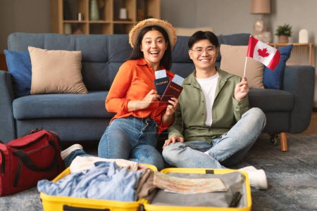 Foto de Emocionada pareja joven asiática posando con bandera de Canadá, empacando una maleta de viaje para un nuevo viaje en casa. Los cónyuges sonriendo a la cámara preparándose para la reubicación y viaje al extranjero - Imagen libre de derechos