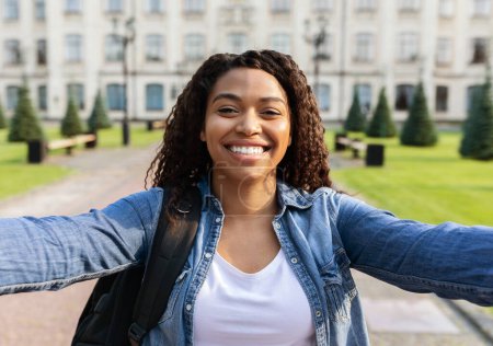 Foto de Primer plano de hermosa joven estudiante tomando selfie al aire libre, mujer feliz usando mochila posando fuera contra edificio de la universidad, mirando y sonriendo a la cámara, espacio libre - Imagen libre de derechos