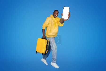 Photo for Glad young african american guy goes with suitcase, freezes in air, shows smartphone with empty screen, isolated on blue studio background. Recommendation app for travel, holidays, weekends - Royalty Free Image