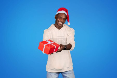 Téléchargez les photos : Souriant jeune homme afro-américain en sweat-shirt, chapeau de Père Noël, tenir boîte cadeau isolé sur fond bleu, studio. Présenter une idée pour Noël, Nouvel An, annonce et offre, célébrer les vacances - en image libre de droit