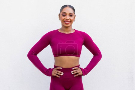 Photo for Sport And Healthy Lifestyle. Fit young black woman exercising over white wall background. Portrait of confident lady training at gym, wearing pink sportswear, hold hands on waist, smiling at camera - Royalty Free Image