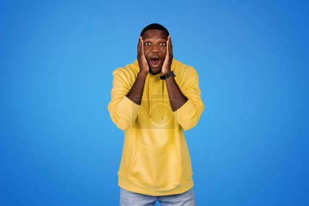 Photo for Glad shocked young african american guy in sweatshirt with open mouth presses hands to face, isolated on blue studio background. Lifestyle, surprise, great idea, ad and offer - Royalty Free Image