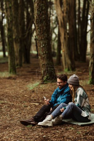 Téléchargez les photos : Positif jeune couple caucasien en vestes dans la forêt, profiter des voyages, vacances assis sur le tapis, utiliser l'application de carte téléphonique, en plein air. Randonnée ensemble, mode de vie, blog d'aventure et de tourisme - en image libre de droit