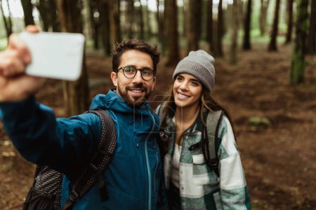 Foto de Alegre joven pareja caucásica en chaquetas en el bosque, hacer selfie en el teléfono inteligente, disfrutar de vacaciones, al aire libre. Blog de viajes y redes sociales para senderismo, estilo de vida, aventura y turismo - Imagen libre de derechos