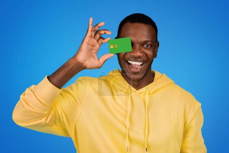 Téléchargez les photos : Joyeux beau jeune homme afro-américain en sweat-shirt presse carte de crédit à l'oeil, s'amuse, isolé sur fond de studio bleu. Vente, finance, shopping et argent, publicité et offre - en image libre de droit