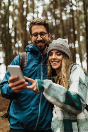 Téléchargez les photos : Joyeux jeune couple caucasien en vestes en forêt profiter des vacances de voyage, utiliser smartphone app map pour itinéraire, extérieur, vertical. Dispositif moderne pour la randonnée, l'aventure et le tourisme - en image libre de droit