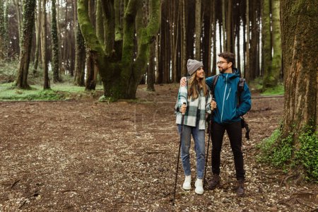Téléchargez les photos : Joyeux jeune couple caucasien en vestes avec des bâtons de trekking marcher dans la forêt ensemble, profiter d'un voyage en plein air, pleine longueur. Randonnée pédestre, mode de vie actif, aventure et tourisme, annonce et offre - en image libre de droit