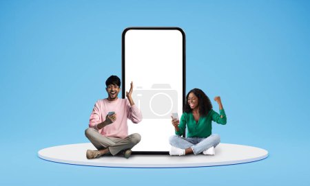 Photo for Great Offer. Happy Black Couple With Cellphones Sitting Near Huge Smartphone With Empty Blank Screen, Excited African American Man And Woman With Mobile Phones Sitting On Blue Background, Mockup - Royalty Free Image