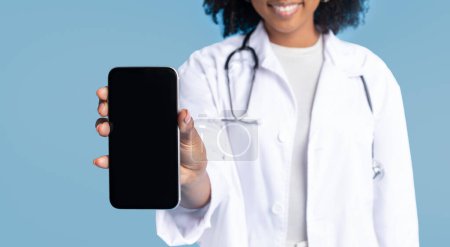 Téléchargez les photos : Souriant noir millénariste dame médecin en manteau blanc montrer smartphone avec écran vide, isolé sur fond de studio bleu, recadré. App pour le travail recommandation professionnelle, médecine et soins de santé - en image libre de droit