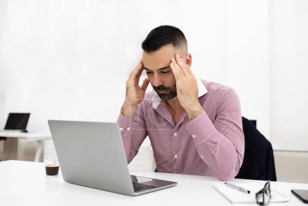 Photo for Unhappy tired caucasian mature guy at table with laptop, presses hands to head, suffers from headache, overwork in coworking office interior. Business problems, work, deadline and stress, pressure - Royalty Free Image
