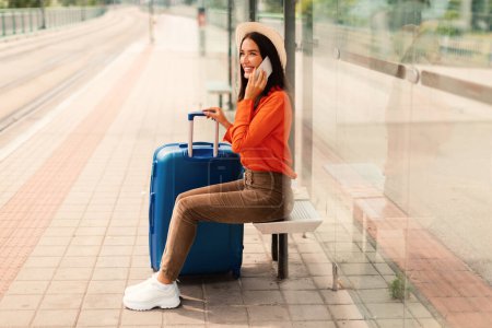 Foto de Mujer pasajera feliz con maleta hablando por teléfono, comunicándose mientras viaja y esperando el tren sentado en la estación de parada de tranvía afuera. Viajes urbanos y comunicación móvil - Imagen libre de derechos