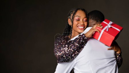 Téléchargez les photos : Ravie jolie jeune femme afro-américaine portant une robe élégante avec Noël présent dans sa main étreignant son petit ami et souriant, couple célébrant le nouvel an 2024 sur fond noir, espace vide - en image libre de droit