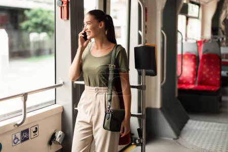 Téléchargez les photos : Toujours en contact. Femme souriante bavardant dans le tramway, navettant engagé dans un appel téléphonique à l'intérieur du train moderne. Passager communiquant et appréciant les transports en commun lorsqu'il se rend au travail - en image libre de droit