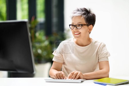 Photo for Cheerful stylish mature woman working from home distance office on pc, typing on keyboard. Smiling 50s middle aged business lady wearing eyeglasses using computer, send emails, chatting with client - Royalty Free Image