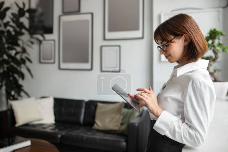 Téléchargez les photos : Jolie jeune femme d'affaires européenne bavardant sur une tablette numérique à l'intérieur du bureau, debout à l'intérieur, espace libre. Affaires, travail et technologie, communication et marketing - en image libre de droit
