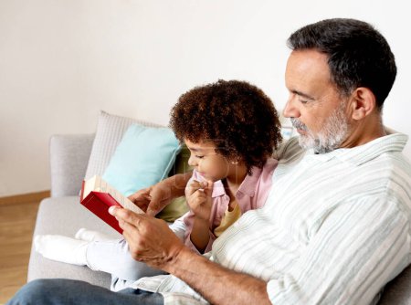 Téléchargez les photos : Joyeux grand-père latin et petit-fils partageant la joie de lire ensemble à l'intérieur de la maison, se détendre et profiter de la littérature, se détendre le week-end, assis sur le canapé à l'intérieur. Focus sélectif - en image libre de droit