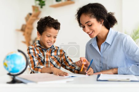 Photo for Kid Education. Tutor Lady Teaching School Boy Sitting At Desk With World Globe Indoors, Explains Lesson. Mom And Son Engaging In Homeschooling, Helping Child Do Homework At Home. Selective Focus - Royalty Free Image