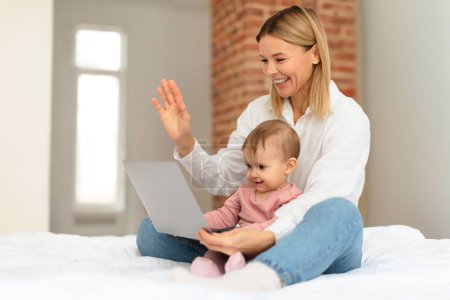 Foto de Feliz madre y su pequeña hija bebé video llamadas en el ordenador portátil, mujer agitando las manos y sonriendo, diciendo hola, sentado en la cama. Comunicación de forma remota, reunión - Imagen libre de derechos