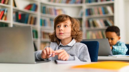 Photo for Concentrated european schoolboy sitting at desk in front of laptop at classroom and listening attentively his teacher, panorama. Modern elementary education - Royalty Free Image