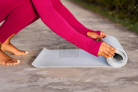 Téléchargez les photos : Recadrée de femme noire en tenue de sport rose déroulant tapis de fitness avant la pratique du yoga en plein air. Athlète féminine se préparant pour l'entraînement de pilates au parc ou au jardin, espace de copie - en image libre de droit