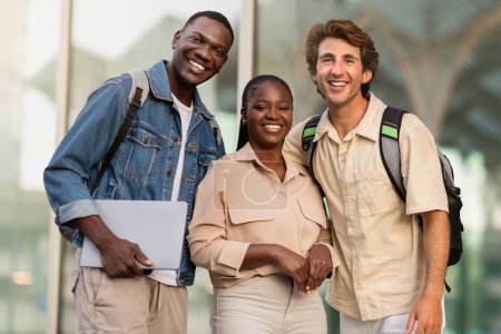 Photo for College Friendship Concept. Happy multiracial students one woman and two guys young friends with backpacks and laptop in hands posing together at university campus, smiling at camera - Royalty Free Image