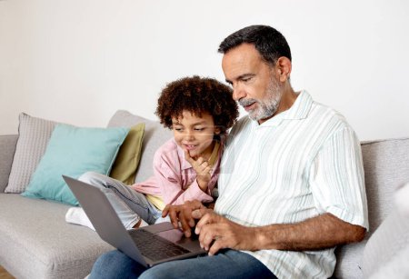 Téléchargez les photos : Grand-père hispanique et petit-fils utilisant un ordinateur portable pour le divertissement en ligne, regarder un film ensemble à la maison. Grand-parent et petit-enfant naviguant sur le Web sur ordinateur. Générations unies à travers les gadgets - en image libre de droit