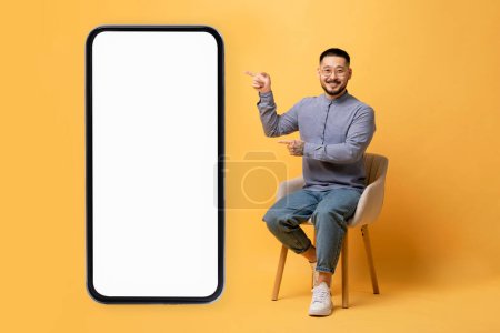 Photo for Positive asian guy pointing at big smartphone with white screen while sitting on chair over yellow studio background, smiling millennial man demonstrating free space for app or website design, mockup - Royalty Free Image