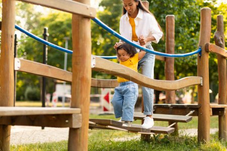 Photo for Korean Mom Guiding Her Baby Daughter Across a Swinging Bridge, Engaging With Toddler in Playground Adventure Fun, Enjoying Outdoor Playtime in Summer City Park. Cropped Shot - Royalty Free Image