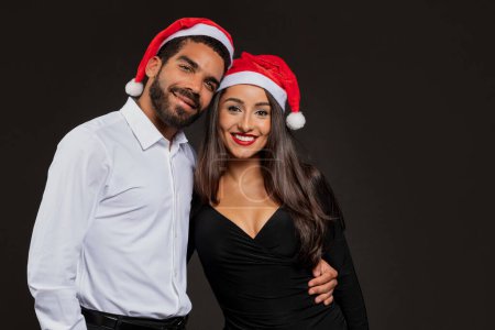 Foto de Hermosa pareja joven hispana feliz en bonitos trajes elegantes y sombreros de santa abrazando y sonriendo a la cámara en el fondo negro, celebrando la Navidad juntos, tienen fiesta de Navidad, espacio vacío - Imagen libre de derechos