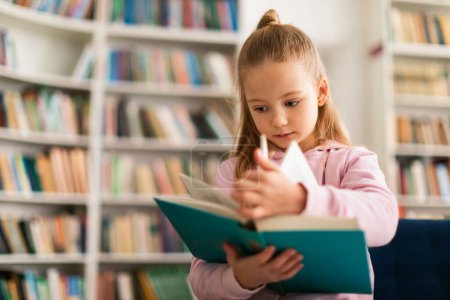 Photo for Cute little school girl leafing through a book, standing in library or classroom at primary school, free space. Girl concentrating to learning by herself. Education and kids concept - Royalty Free Image