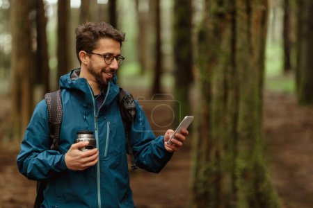 Photo for Positive young caucasian couple in jackets with cup of coffee, walk in forest together, enjoy trip, use phone map, outdoor. Hiking, lifestyle, adventure and tourism alone with device - Royalty Free Image